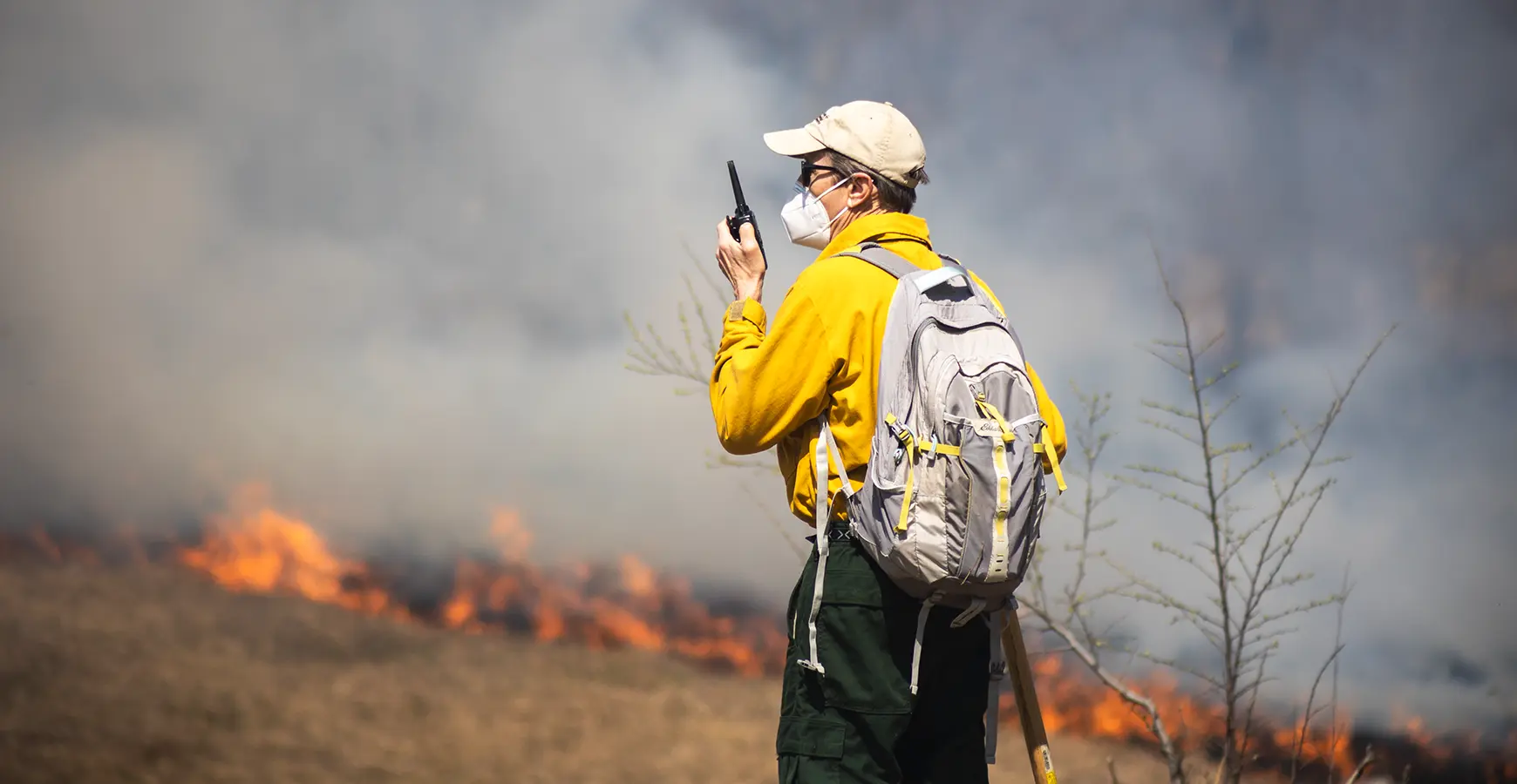 Biology professor and director of Green Oaks, Stuart Alison, watches the prairie burn at Green Oaks. He is dressed in prairie gear: yellow shirt, dark pants, mask, and hat.