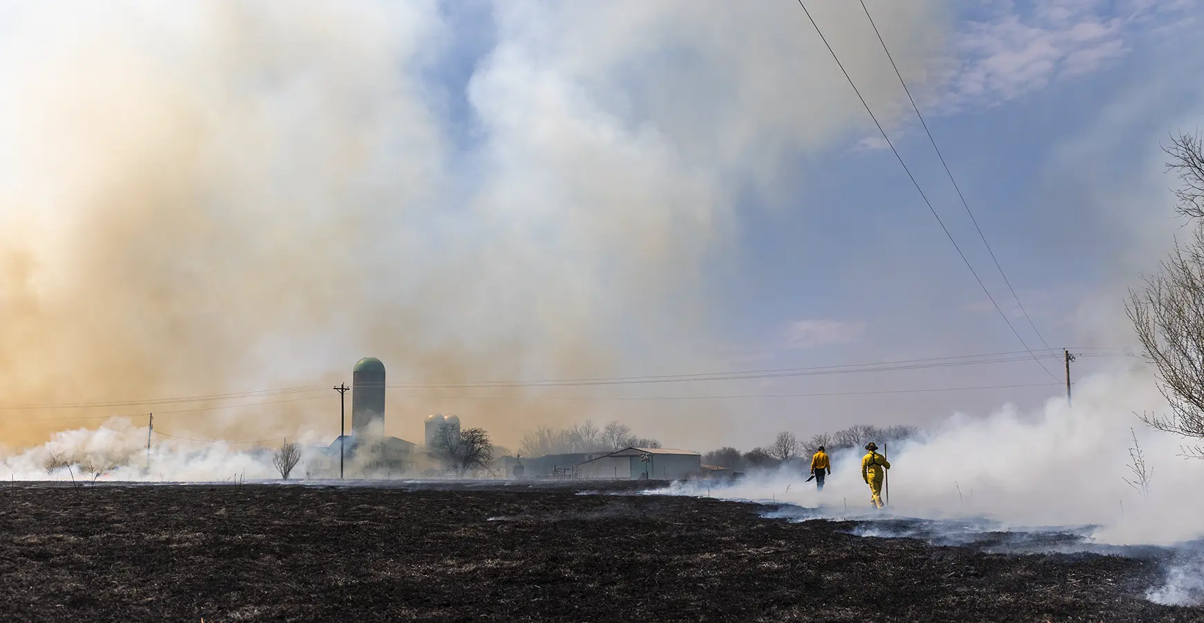 Two Knox students walk the prairie after a prairie burn. They are surrounding by smoke, and a farm scape is in the background.