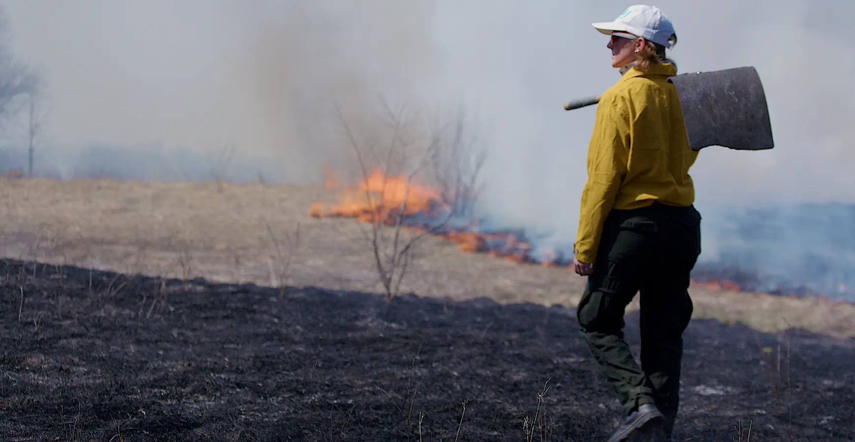 A student in fire gear holds a tool over their shoulder and watches the prairie fire and smoke.