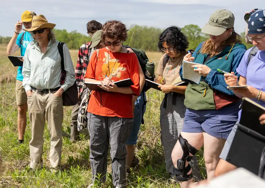 Students stand in a line in a prairie, making notes in notebooks.