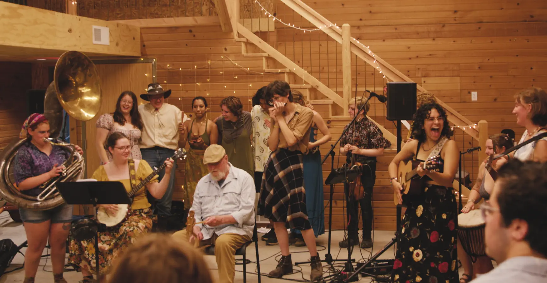In a barn strung with white lights, a group plays musical instruments. Behind them, six individuals link arms.