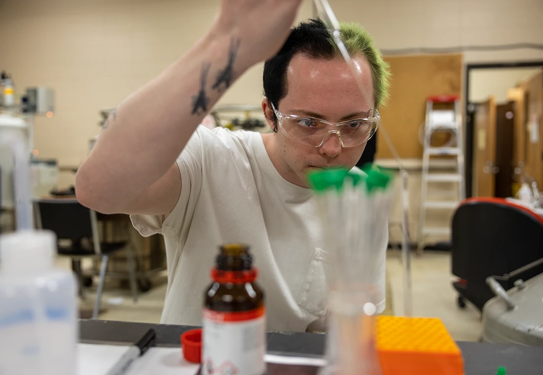 A student wearing goggles carefully adds liquid from a pipette into a beaker.