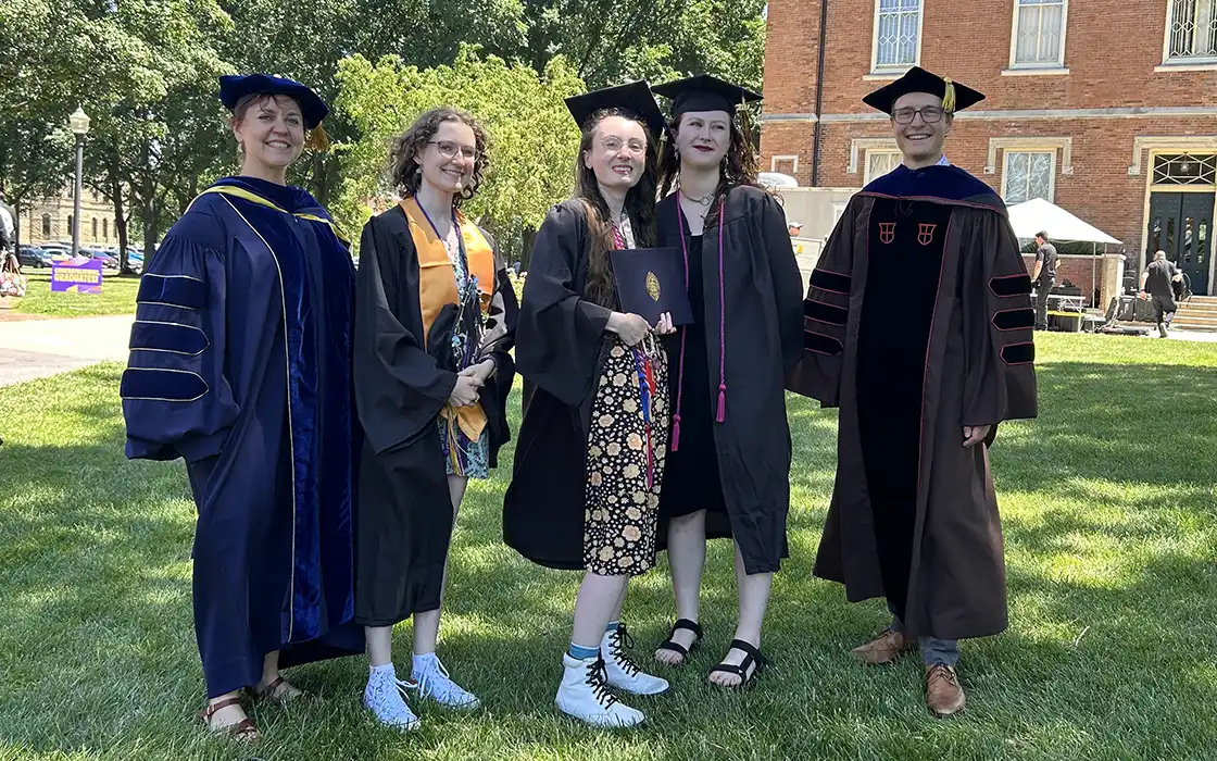 Graduates and professors stand on the lawn in front of Old Main in academic regalia, smiling.