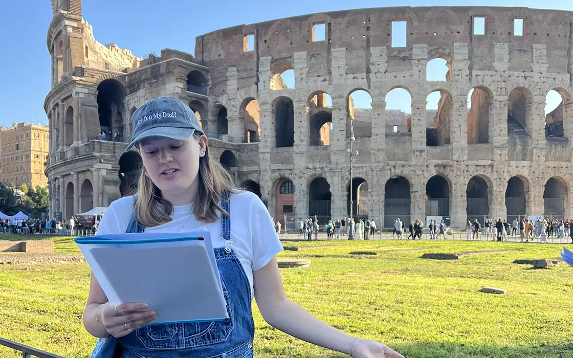 A student stands in front of the Colosseum, gesturing as she reads from a paper.