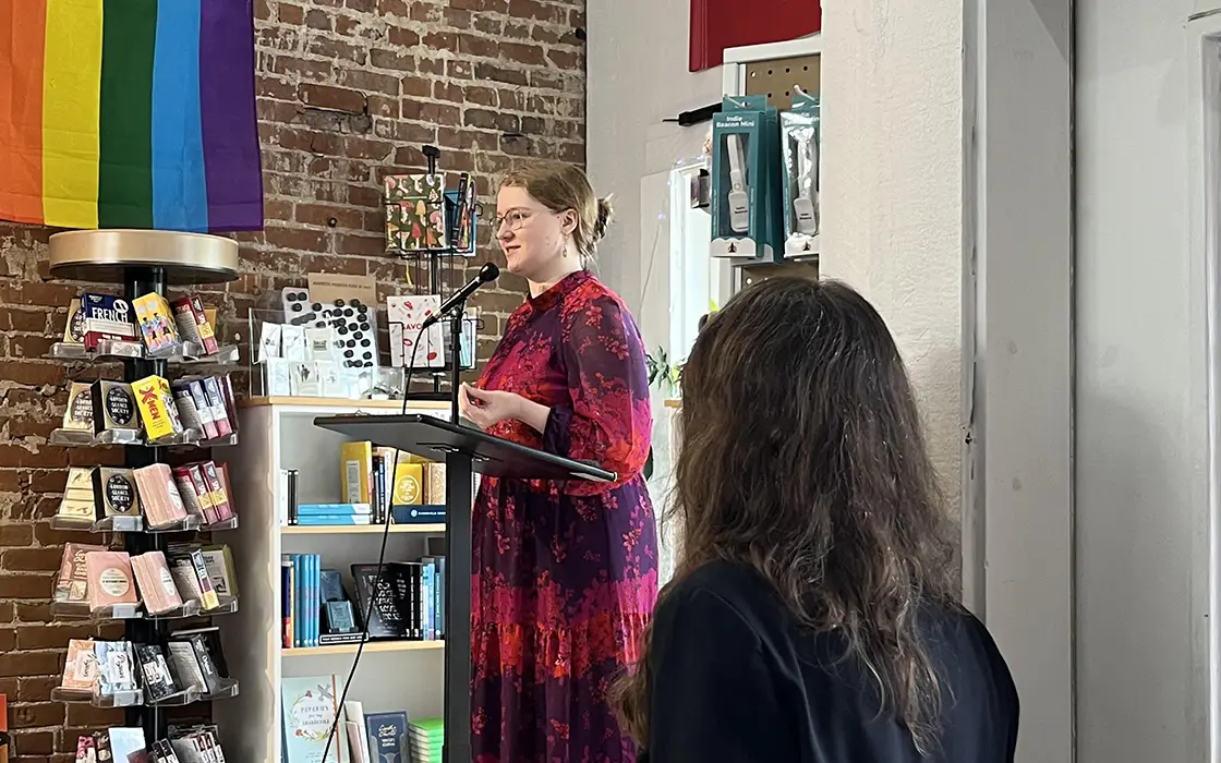 A woman in a maroon dress stands at a podium and speaks. Displays of books are visible in the background.