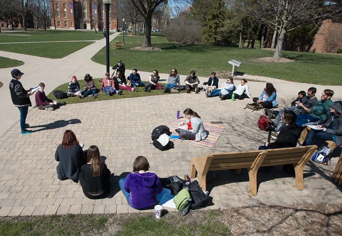 A class sits outside with Old Main in the background as students perform a reading of Homer's classics.