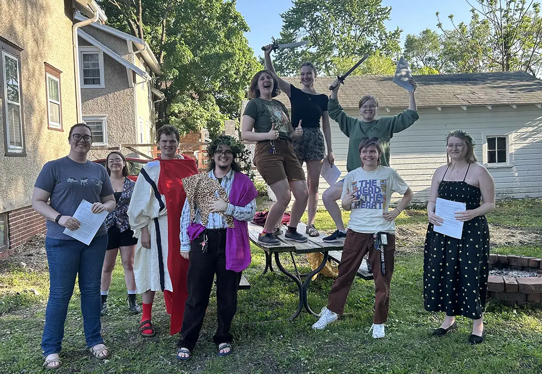 Students gather in a group, some dressed in togas, some standing on a picnic table with plastic swords, smiling at the camera.