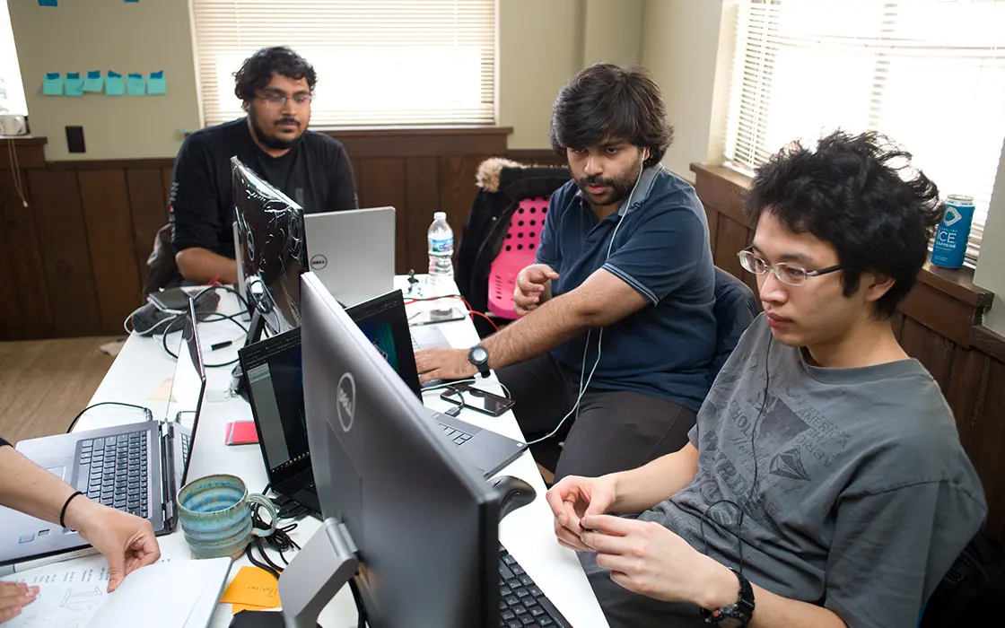 Three students work together with several computers on a table. All students' focus is directed to one student's screen.