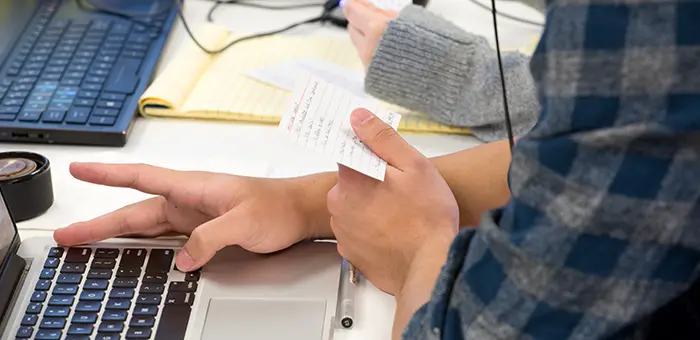 A student's hand is poised over their keyboard. The other hand holds a notecard with handwritten notes.