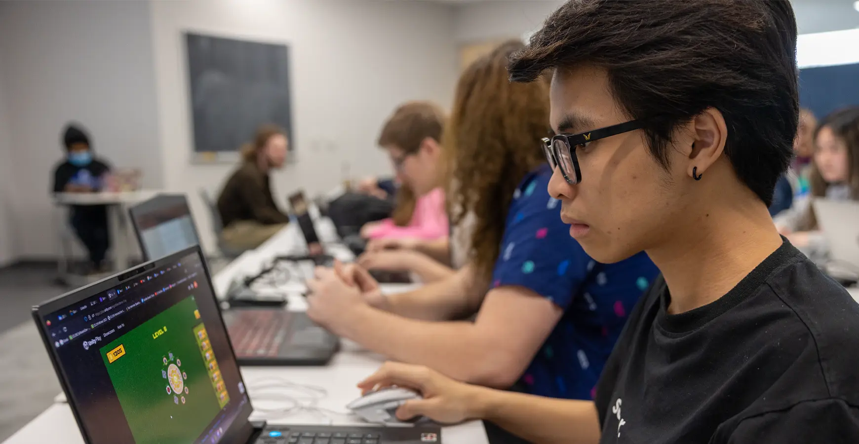 A student works on a laptop. On his screen, a green background is visible with a round graphic and words Level 6 at the top.