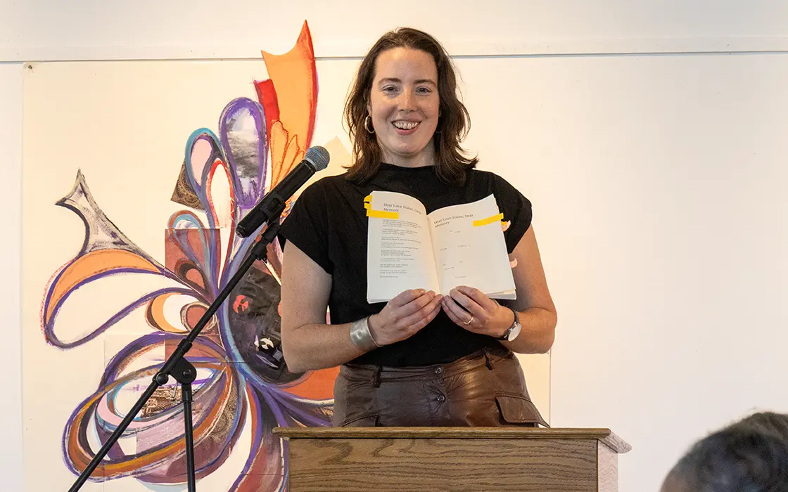 A smiling woman stands at a podium and holds a copy of her book open at the Knox is Lit alumni program.