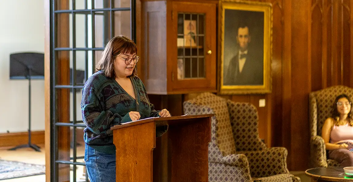 A student stands in front of a wooden podium reading from a manuscript.