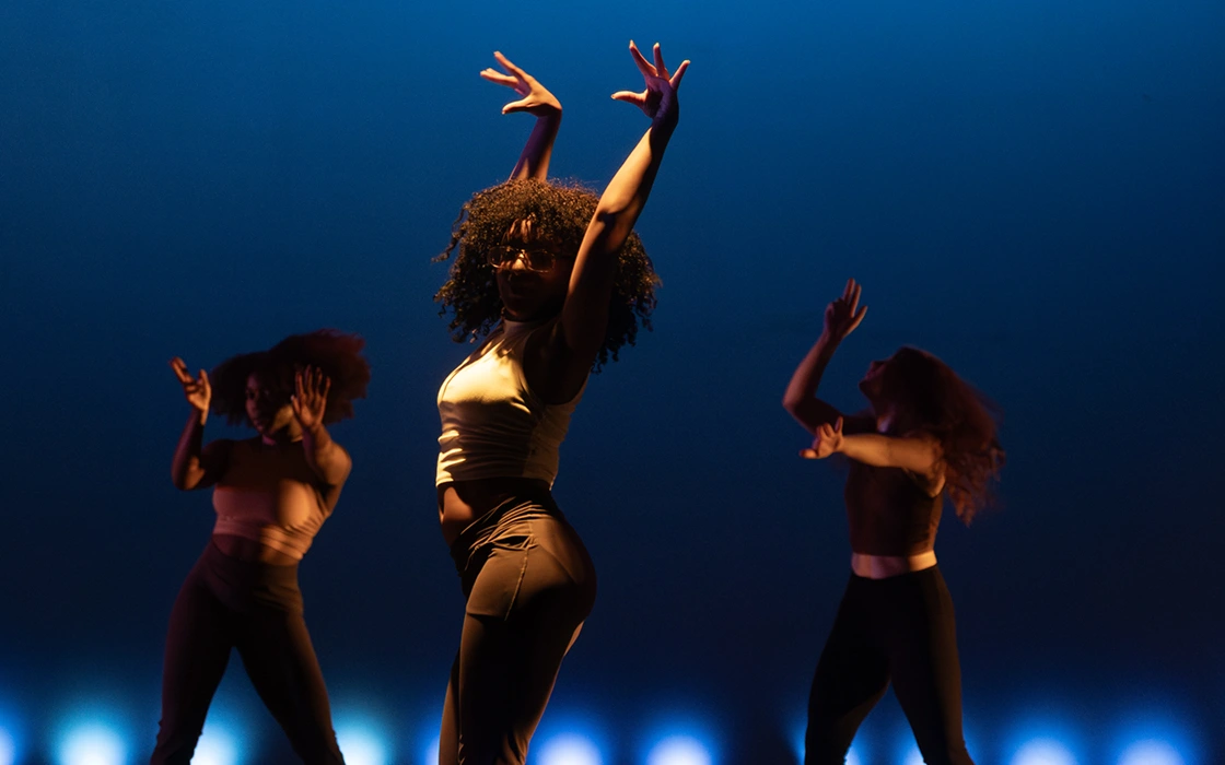 A dancer hits a dramatic pose on stage. Two other dancers are visible in the background. Lighting on stage gives a dark blue background.