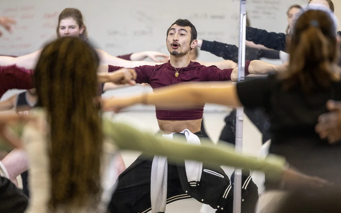 A dance instructor pliés while shouting instruction; students surround the instructor, mimicking his pose.