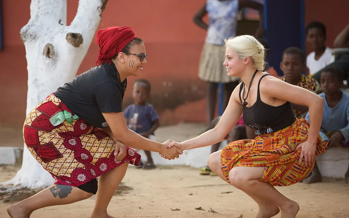 Two students wearing traditional kangas as skirts squat low, facing each other and shaking hands.
