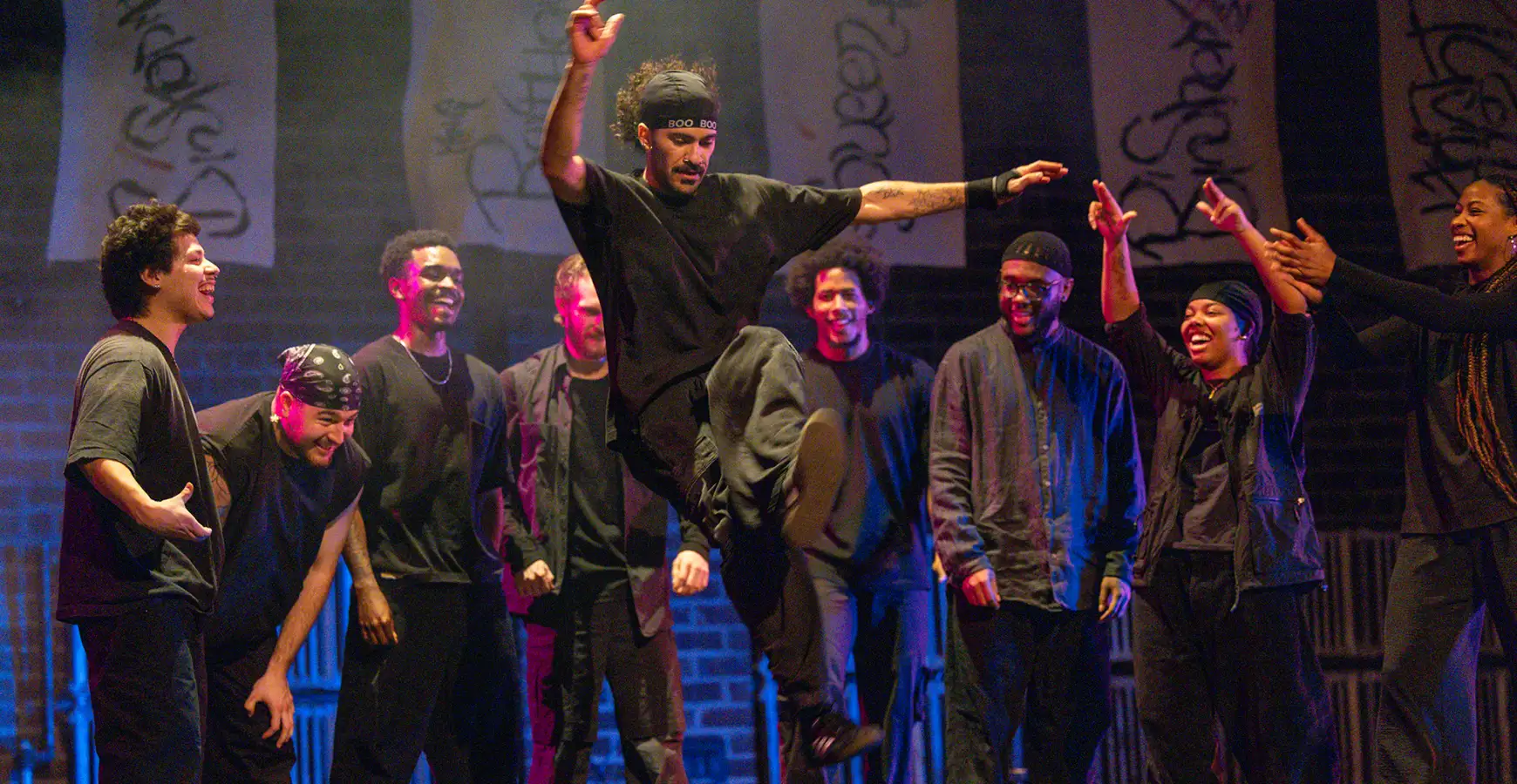 A dancer jumps in the air as he does a trick; eight dancers stand behind him, smiling and cheering him on.