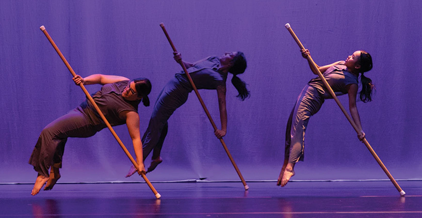 On a stage with deep purple background lighting, three dancers are caught mid-move, suspended above the stage with a long stick.