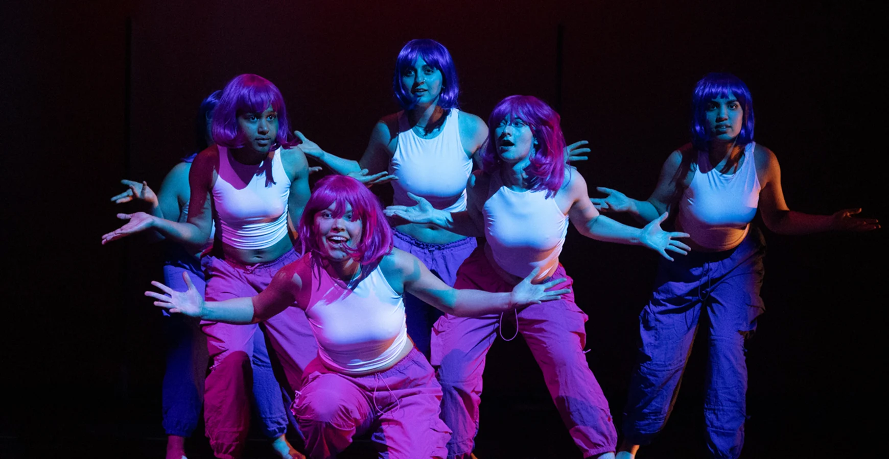 During a performance, five dancers wearing pink and purple wigs, dressed alike, pose with their hands out.