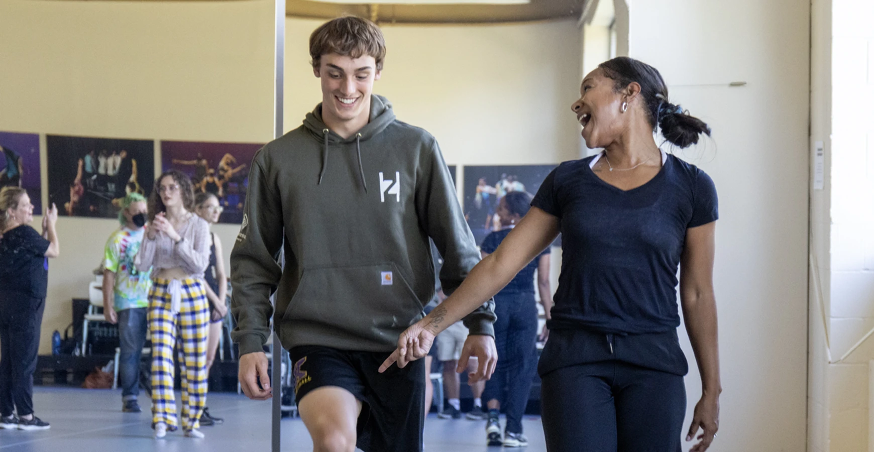 In a dance studio, two dancers smile as one points to the other's knee. More dancers are visible in the reflection of the mirror behind them.