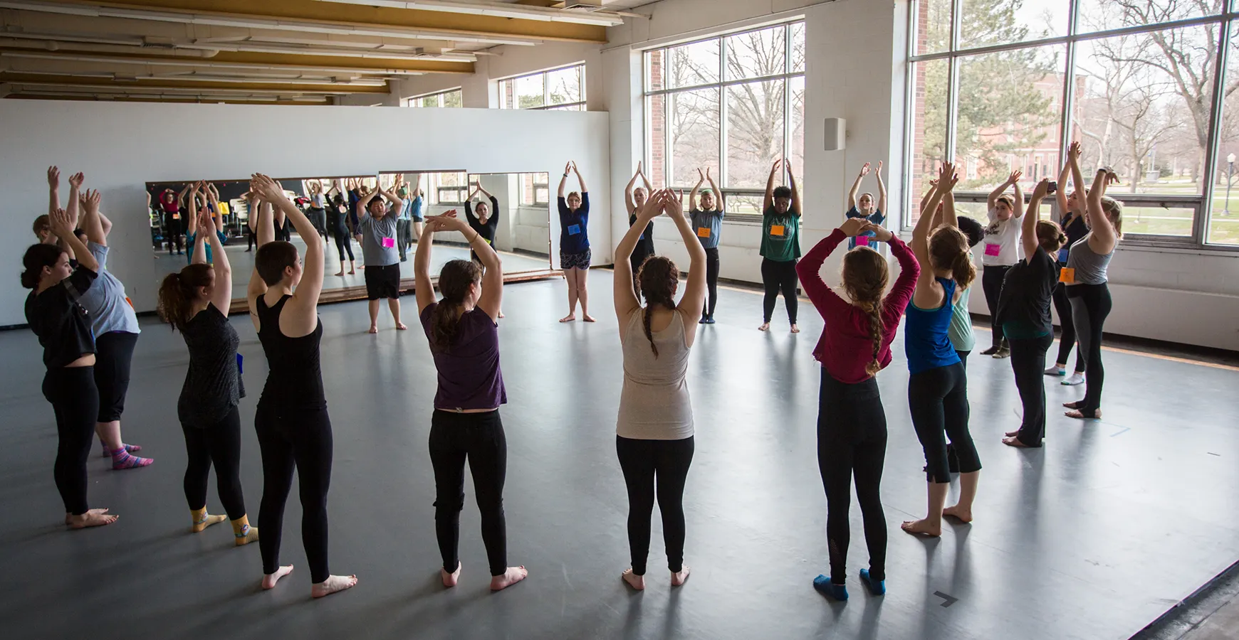 In a dance studio with large windows looking out into campus, 20+ dancers stand in a circle with their arms up in a stretch position.