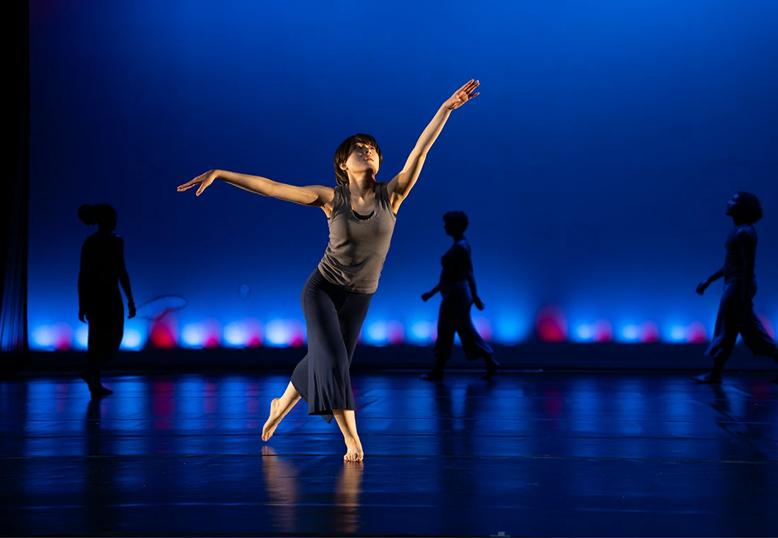 A dancer strikes a pose on stage with blue lights in the background. Silhouettes of three other dancers are also visible in the background. 