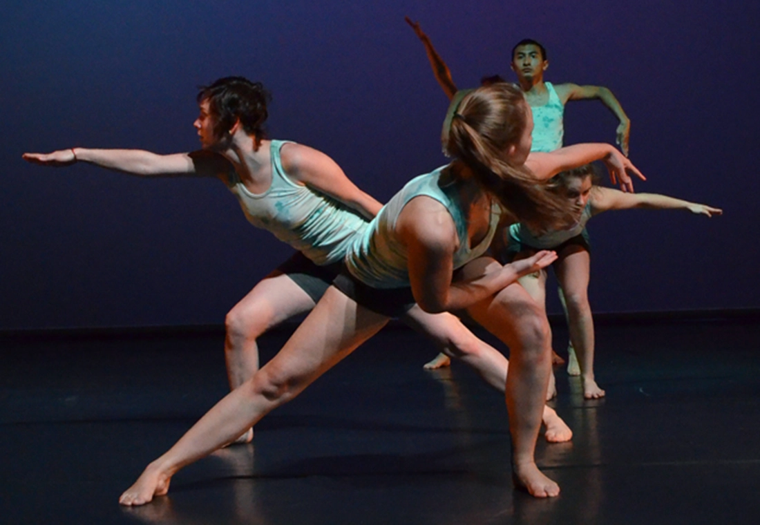 Dancers in a line strike poses on stage with dark blue lighting around them.