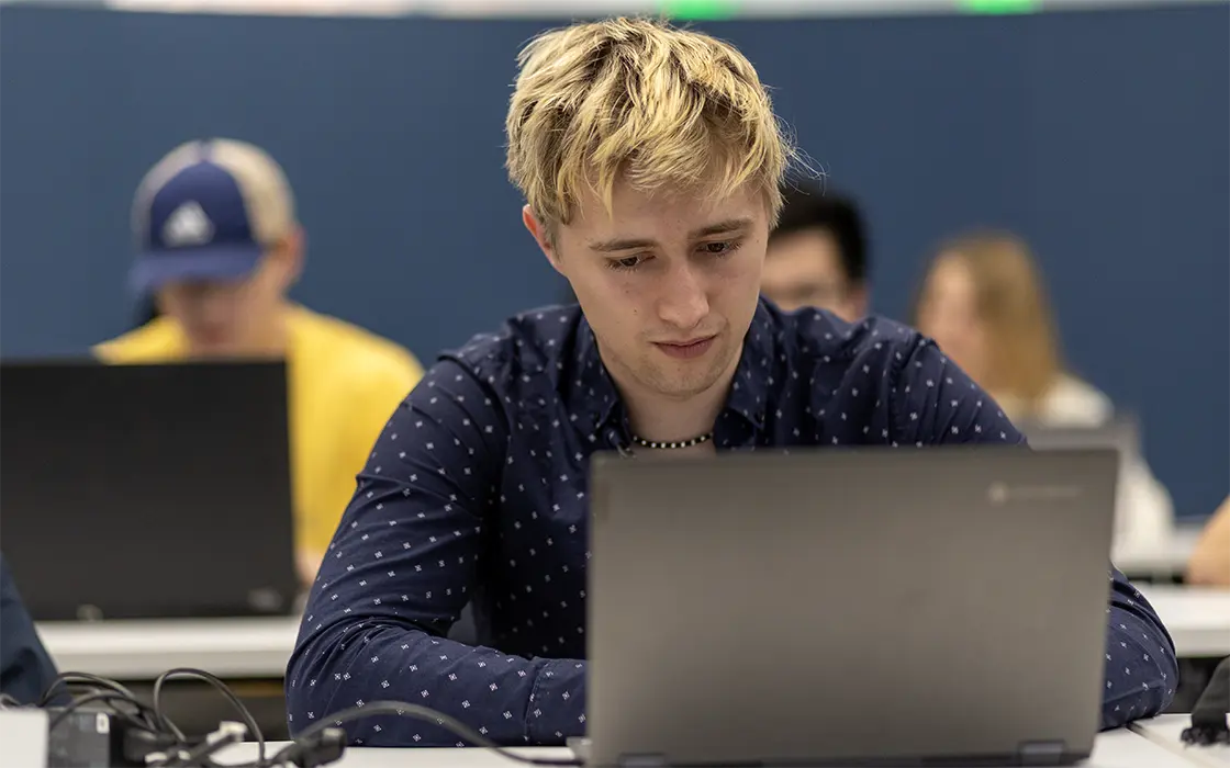 A student works on a laptop. More students on laptops are visible in the background.