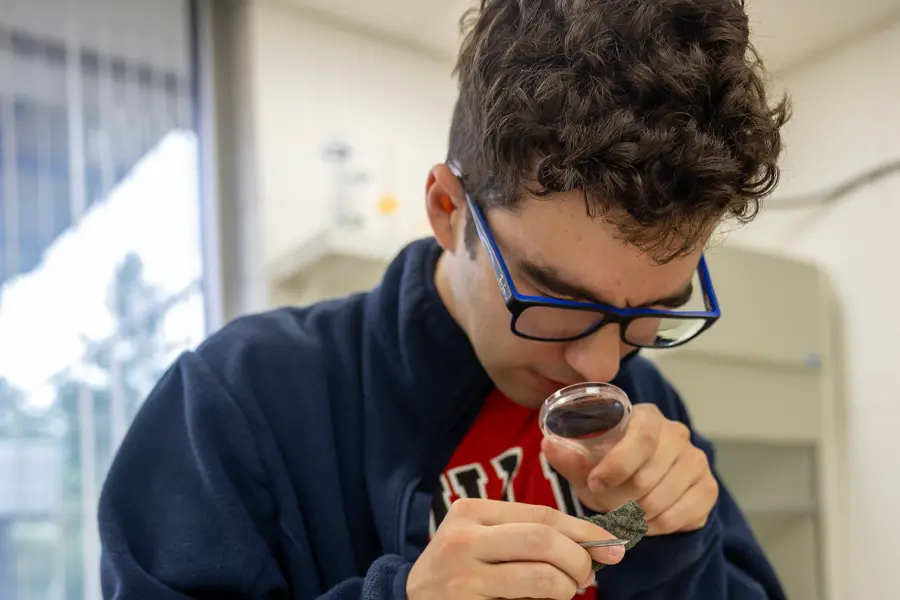 A student squints one eye and looks through a magnifying glass, examining a rock.