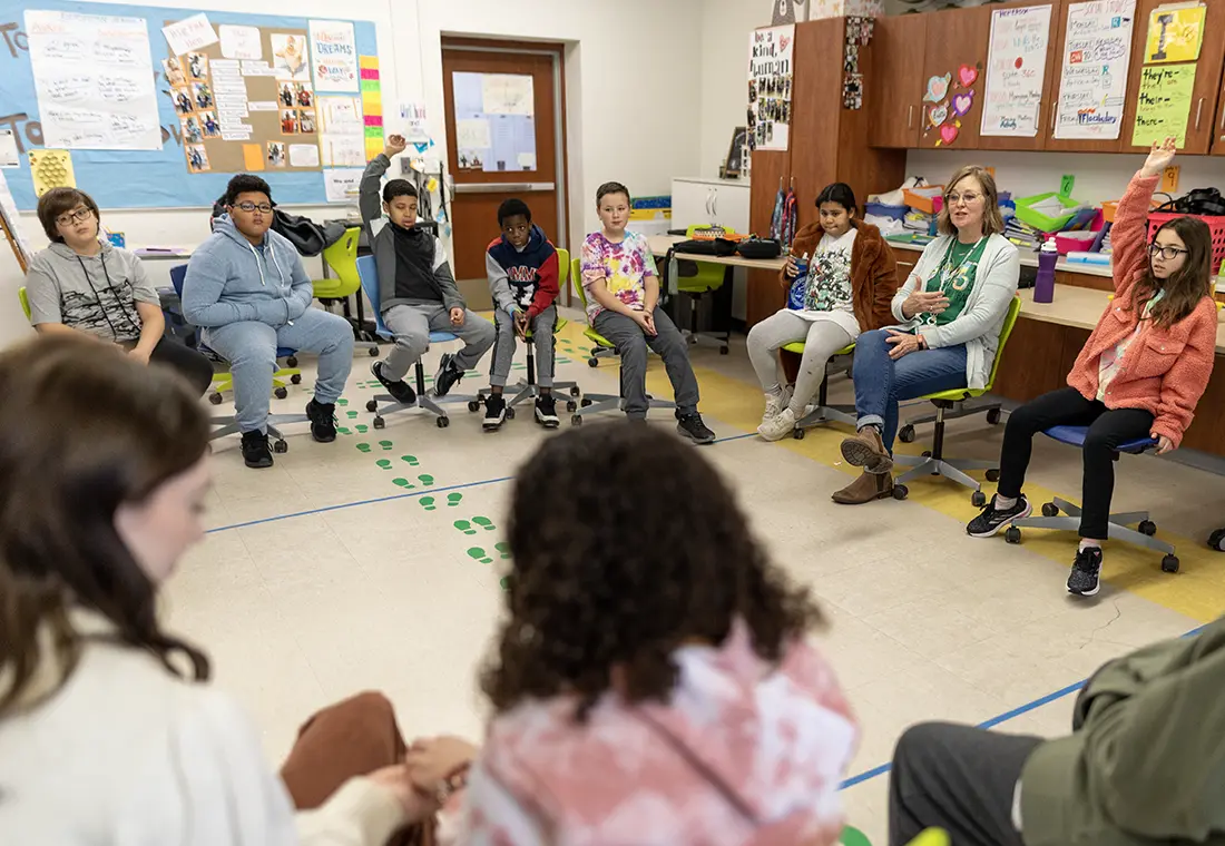 Elementary students sit in a circle. A teacher leads discussion, and two students raise their hands.