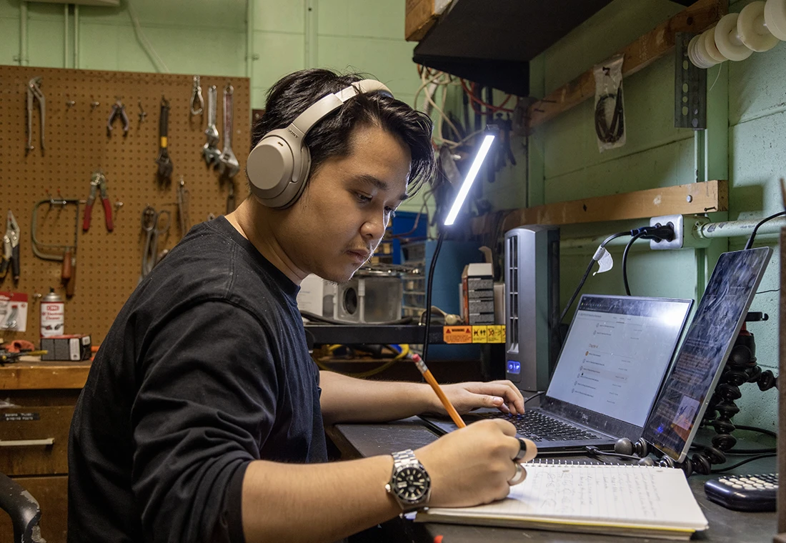 A student makes notes while working on a laptop. He is wearing headphones and working in a shop environment.  