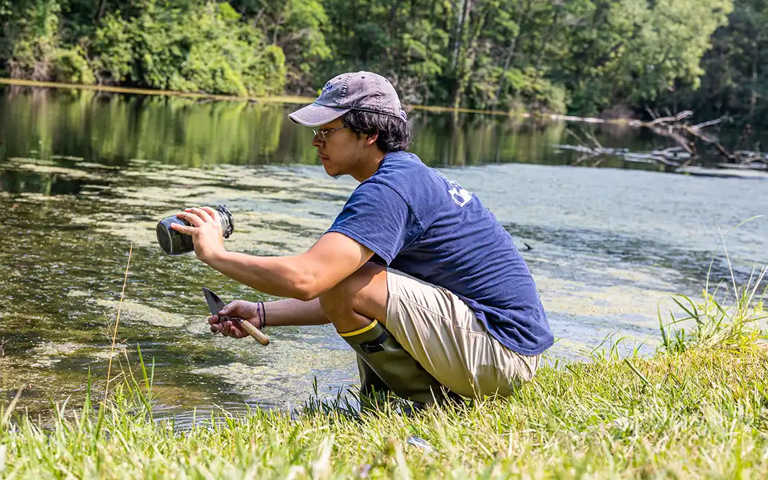 A student wearing rubber boots squats next to a body of water with a spade in his hand, collecting a water and soil sample.