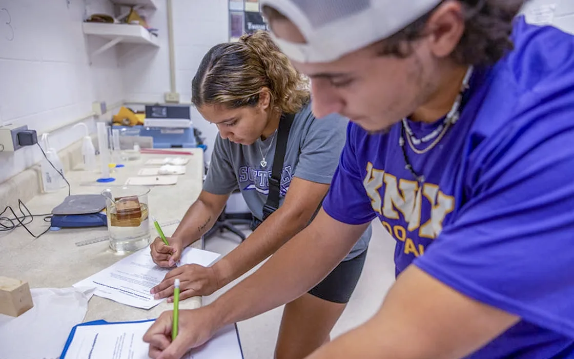 Two students work on a lab, writing notes while a solution sits on the counter.