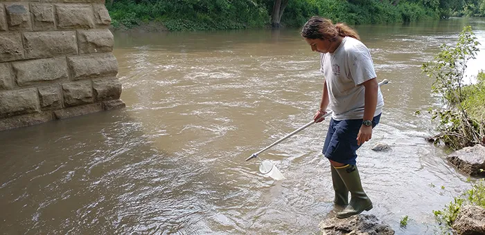 A student wearing rubber boots walks in a body of murky water holding a sample collection tool.