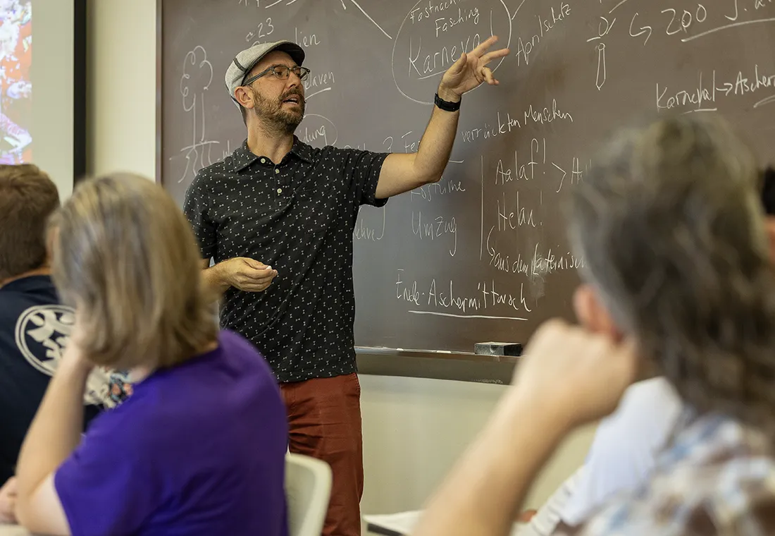 A professor points at a chalkboard. German words are on the board. Students are visible in the foreground looking at the board.
