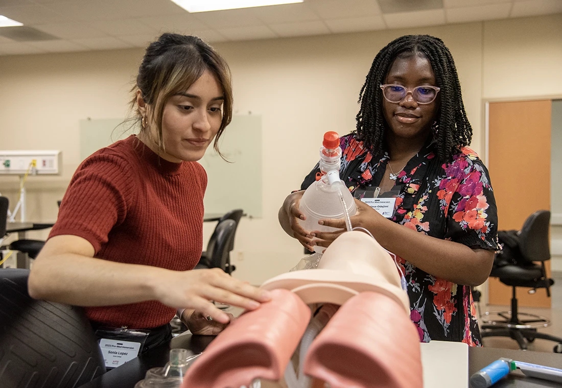 Two students work on a medical manikin. One holds a plastic jug with a tube attached that feeds into the manikin. 