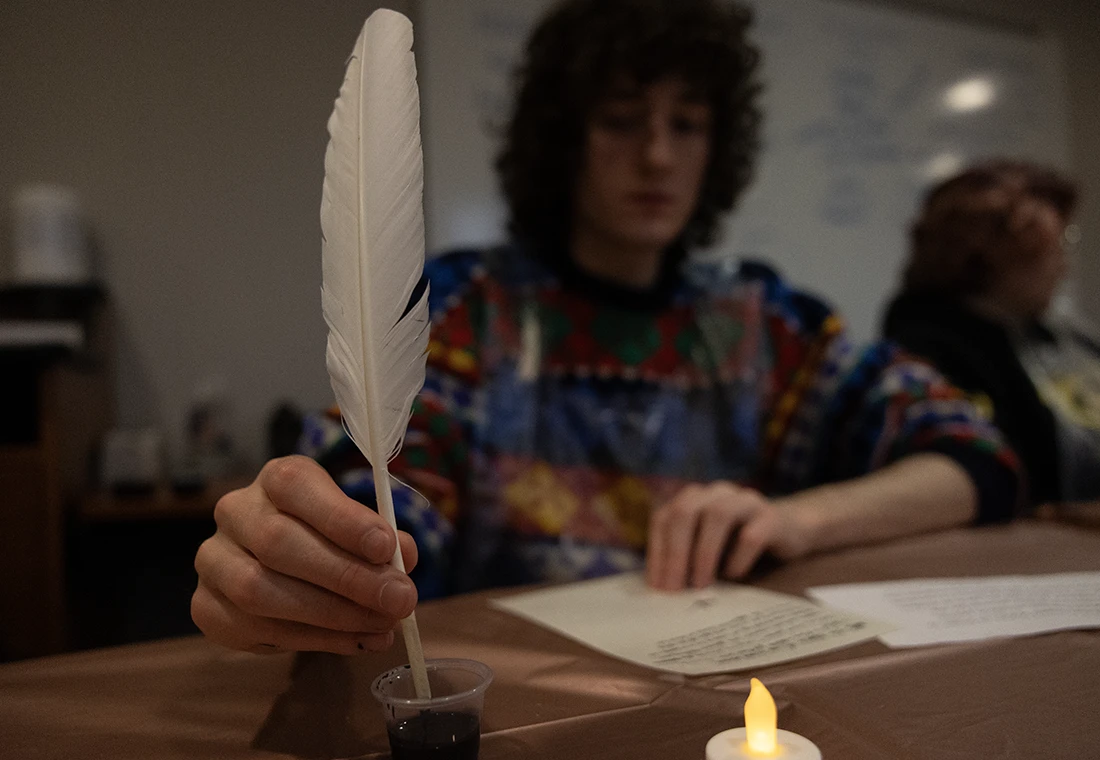 A student dips an old-fashioned quill into a pot of ink. The focus is on the quill; the student is blurred.
