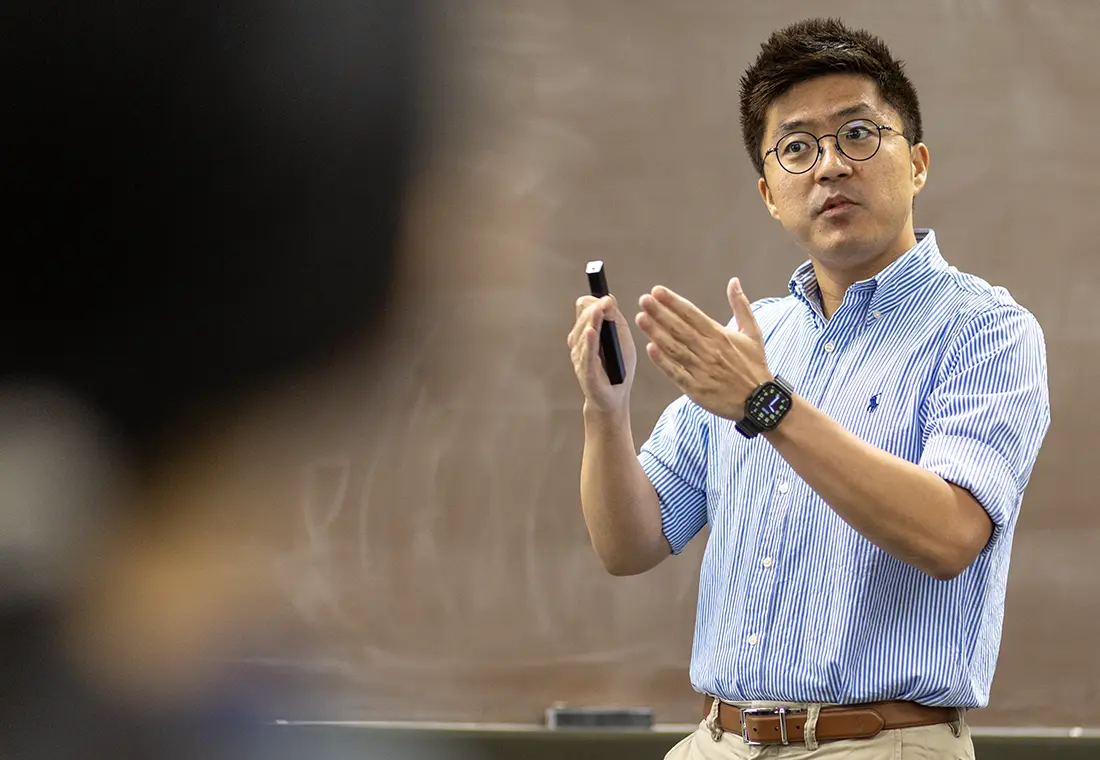A man gestures while speaking. A chalkboard is visible behind him, and a student is visible in front of him.