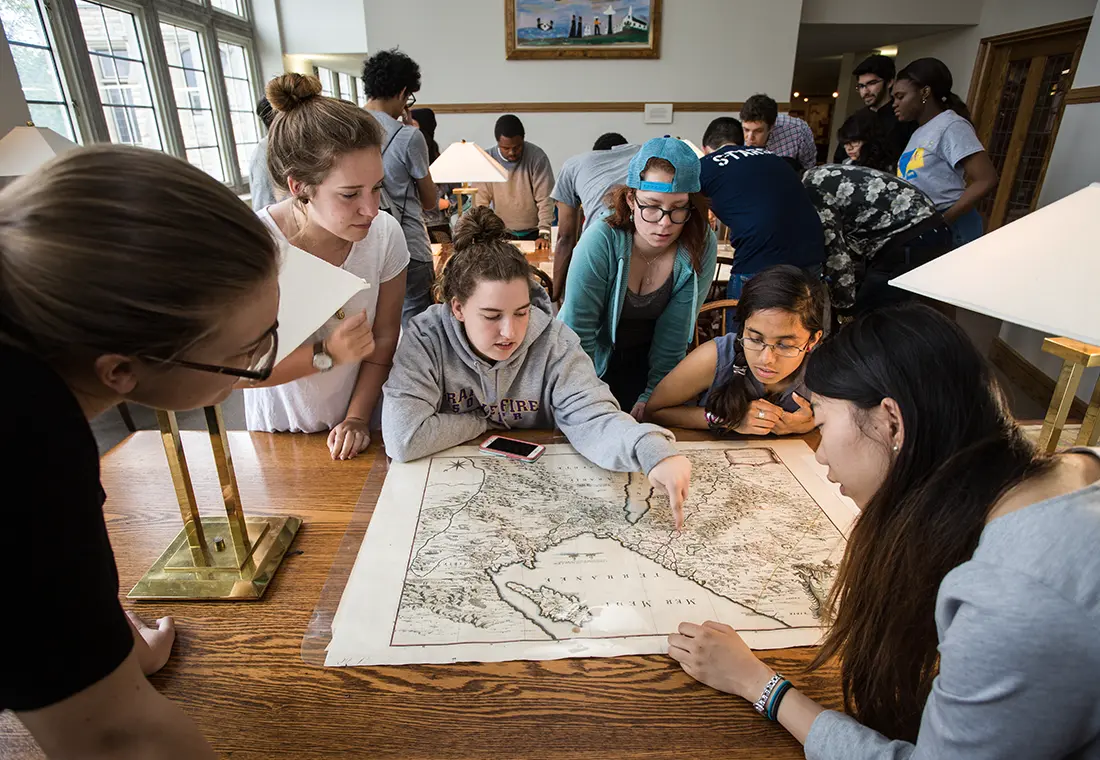 A group of six students looks at a map together, and one points at the map. Another group of students is visible behind them.