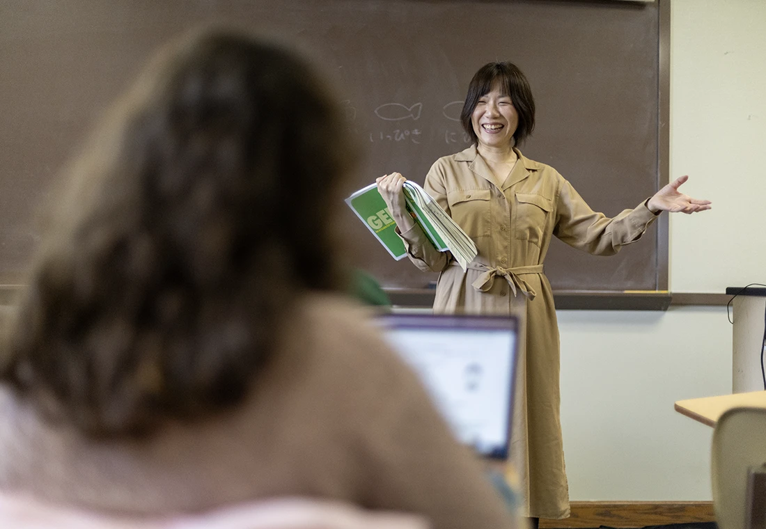 A woman stands in front of a chalkboard with a book in her arm. She is turned toward the classroom, smiling.