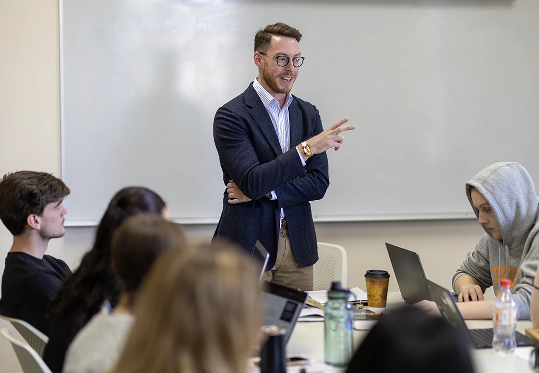 Professor Thomas Bell stands in front of a table, smiling as he speaks. A whiteboard is behind him, and he's surrounded by students who sit at the table.