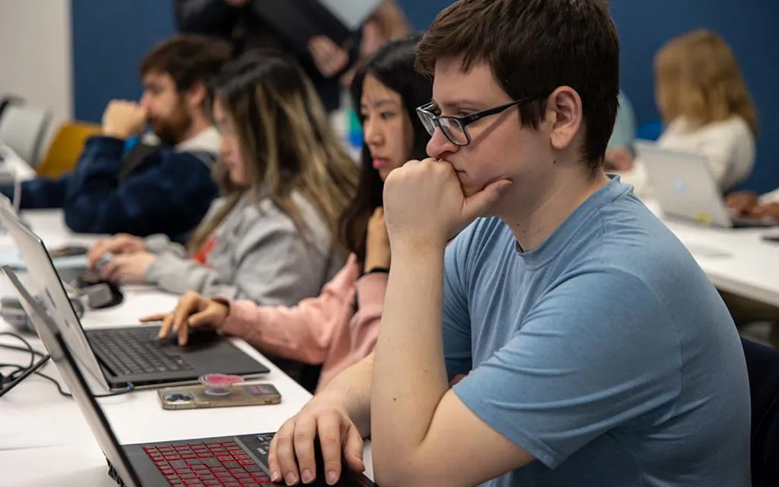 A student working on a laptop stares at the screen, his left hand covers his mouth with a fist as he thinks. Behind him, several other students are working on laptops.
