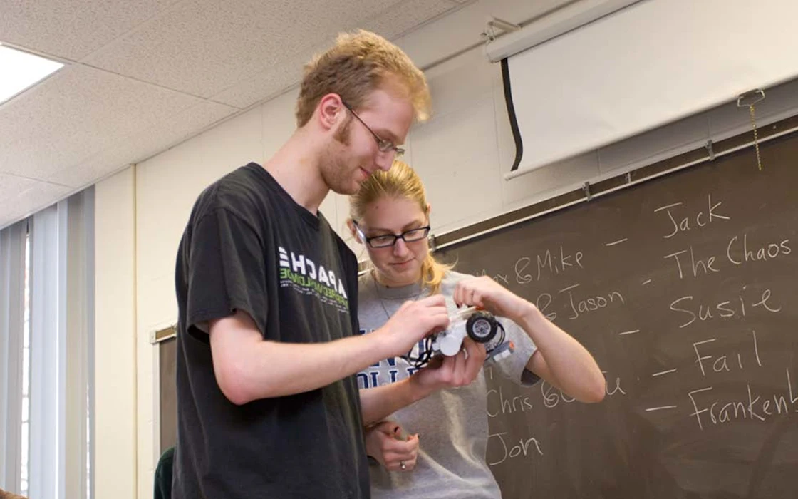 Two students hold a white object with a black wheel. They look down at the object with slight smiles. A chalkboard is behind them.