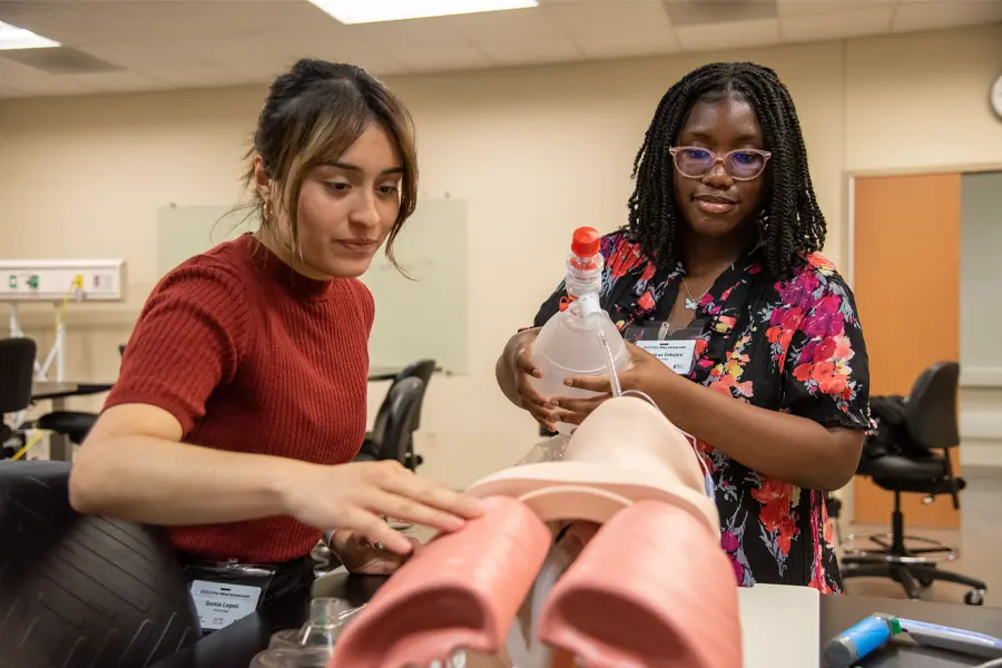 Two students work on a medical manikin. One holds a plastic jug with a tube attached that feeds into the manikin. 