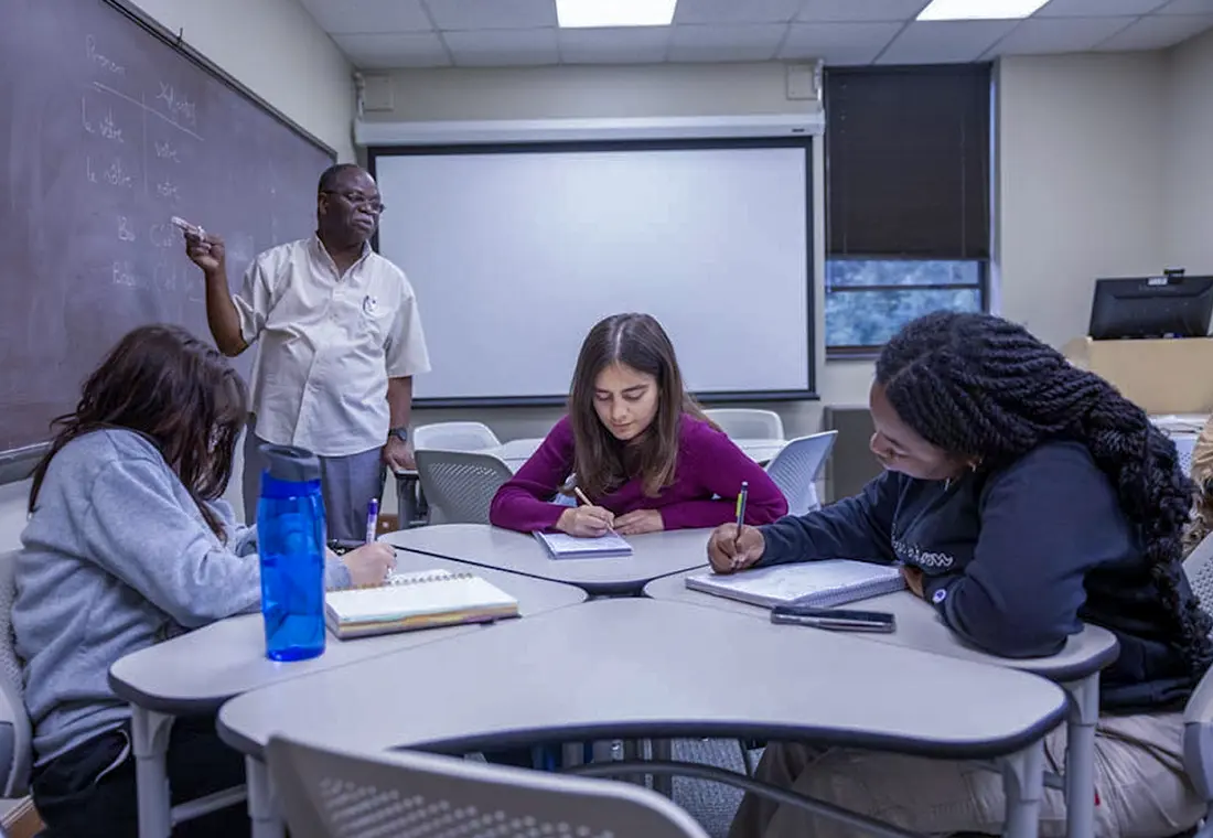 A professor and a student looks at the student's paper together. Other students are visible in the foreground and background. 