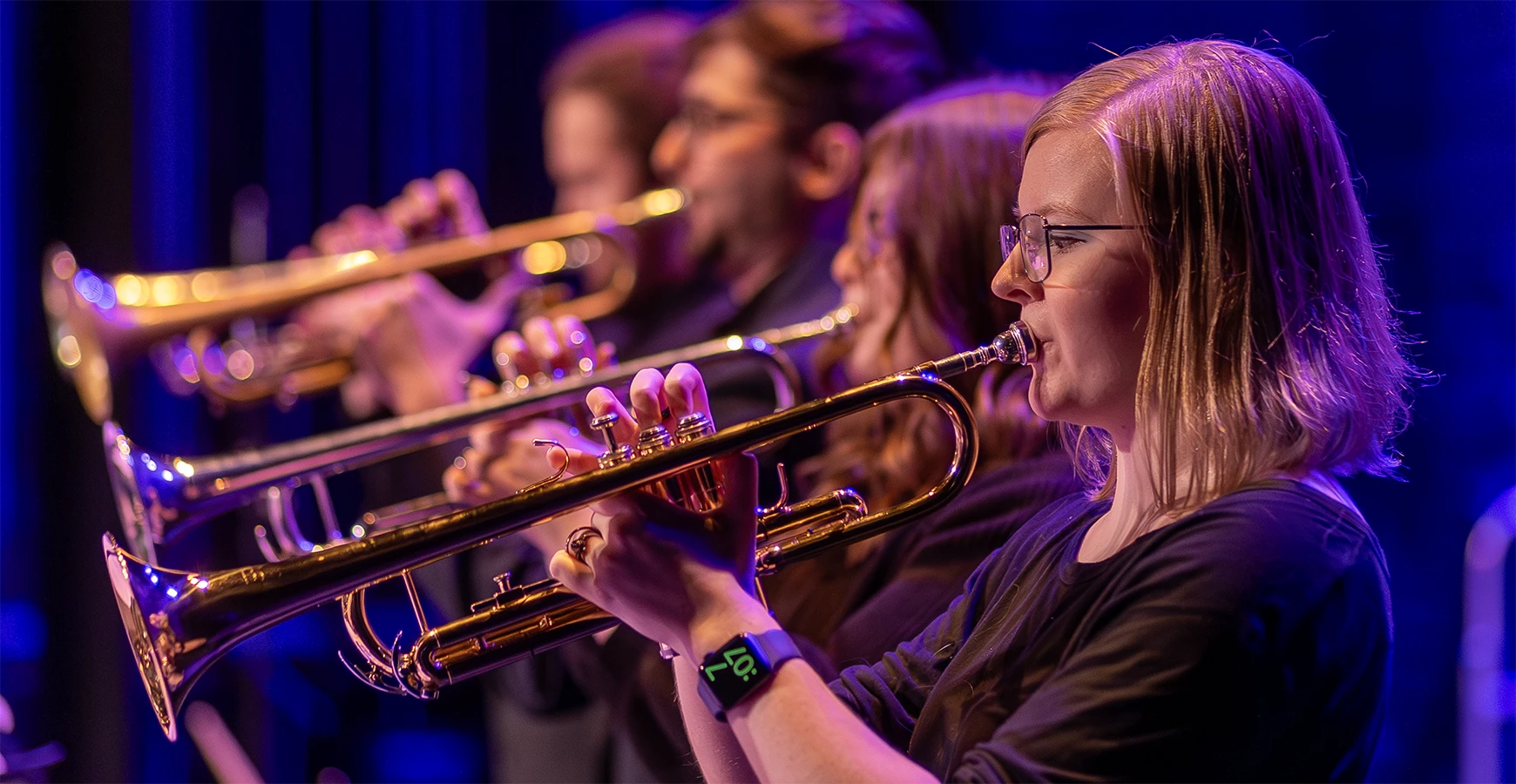 Four trumpeters play  their instruments during a concert; they are framed by purple lights.