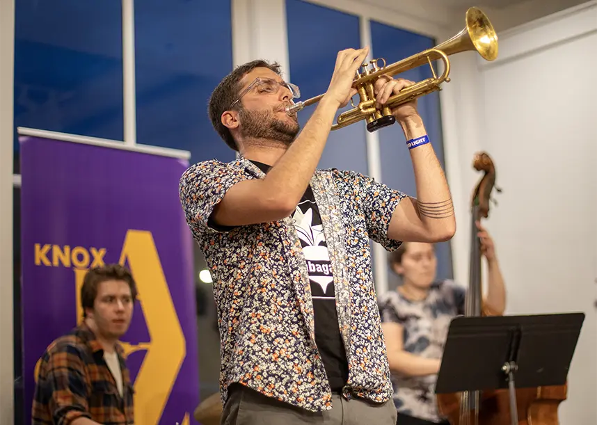 A trumpeter plays in front of two windows at Jazz night. He is joined by a drummer and a bassist.