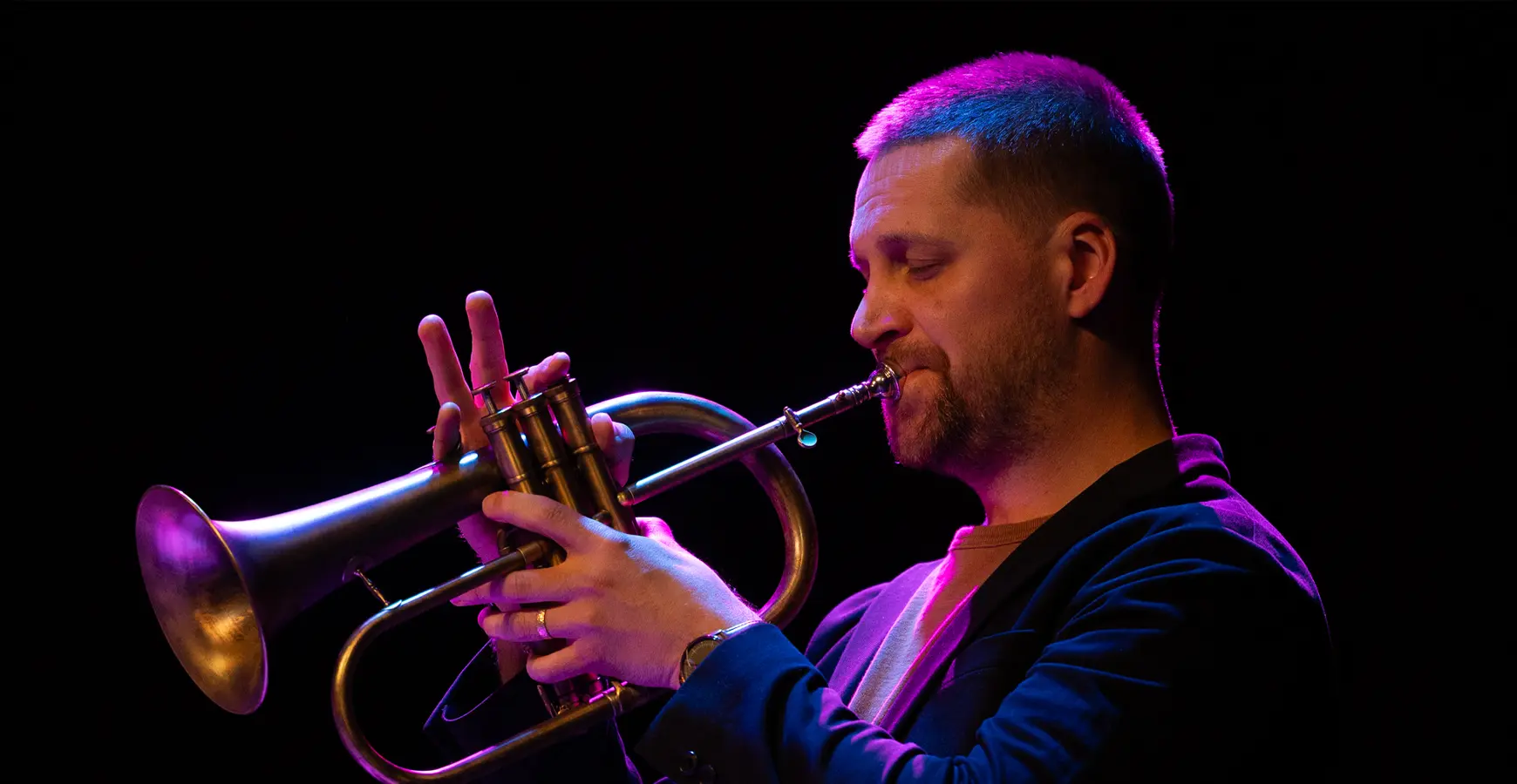 A man plays the trumpet. The background is completely black, and he's cast in a purple hue from stage lighting.