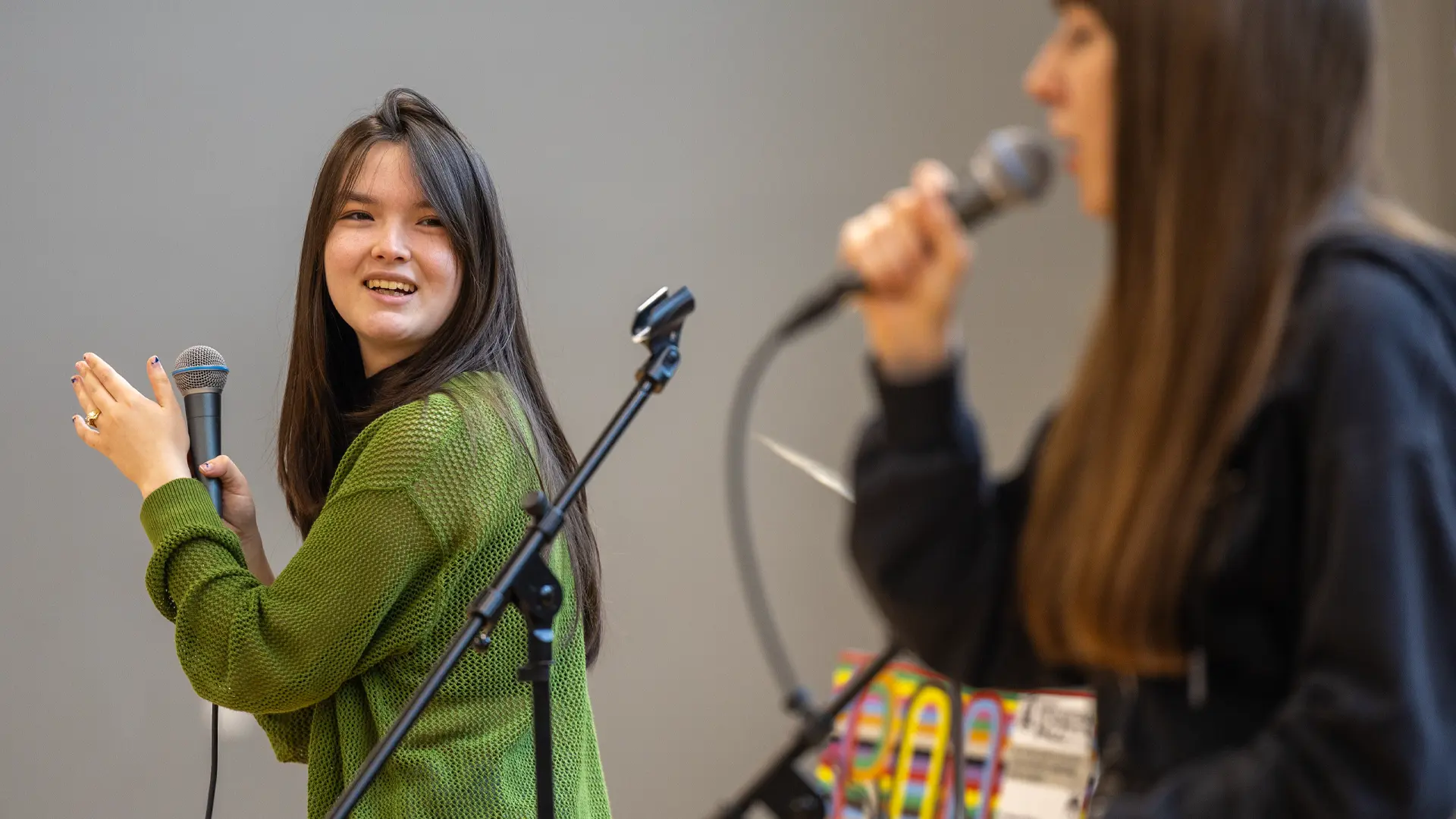 A student holds a microphone and smiles as she glances back at another student who is singing.