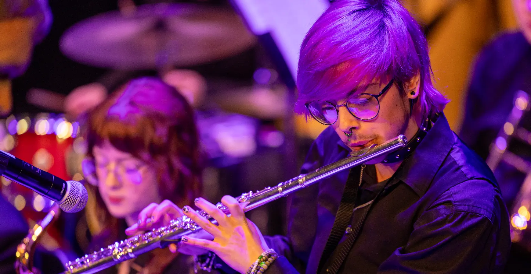A performer plays a flute in front of a microphone; purple stage lighting casts a purple glow.