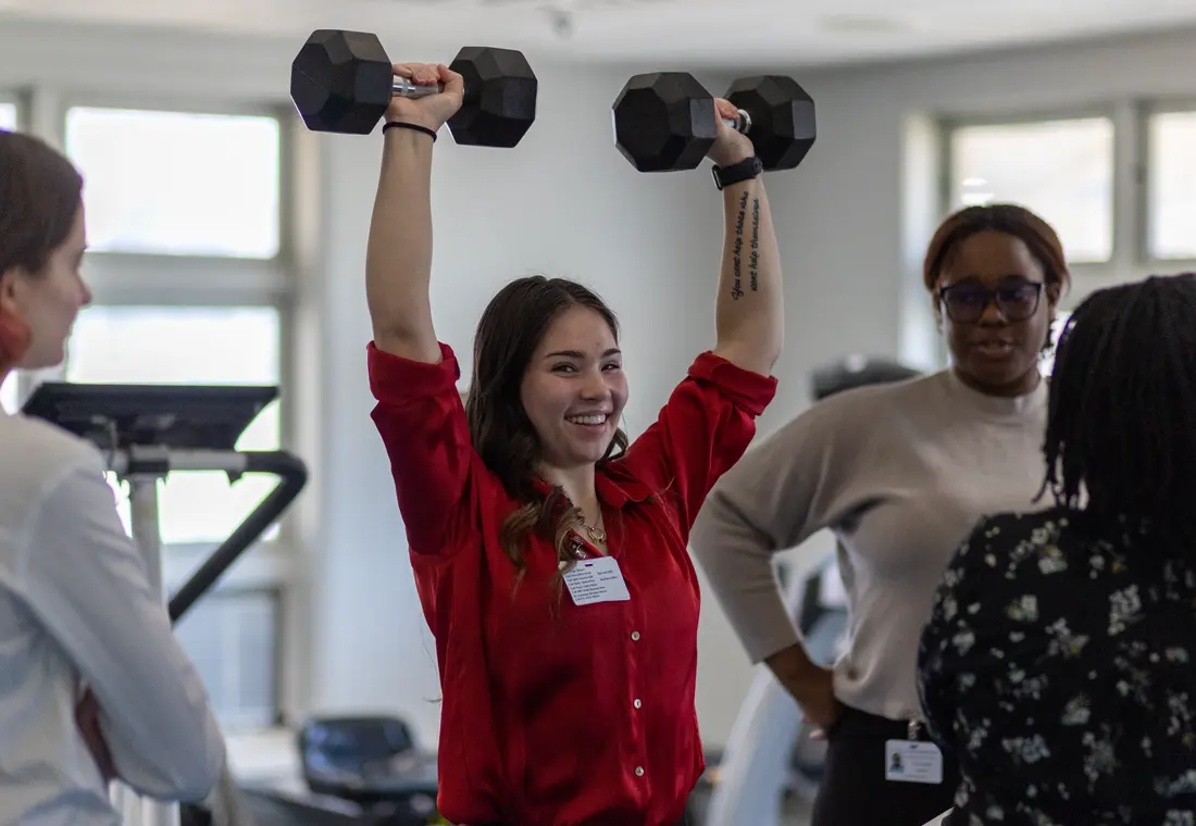 In a gym environment, a student in a red shirt lifts dumbbells over her head, smiling at the camera. Two other individuals stand near her.